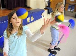 Two girls juggle balls and scarves indoors, one in the foreground concentrating on tossing a yellow and blue ball.