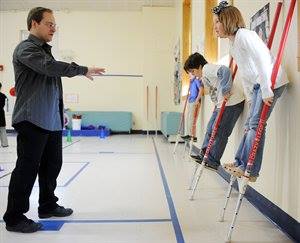 An instructor guides two children balancing on stilts in a gymnasium.