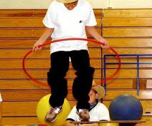 A person wearing a straw hat jumps through a red hula hoop in a gym with wooden bleachers in the background.