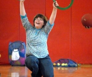 Person kneeling on a gym floor, holding two rings overhead with a red ball and equipment in the background.