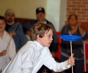 A boy in a white shirt balances a blue plate on a stick indoors, with several people watching in the background.