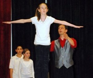 A girl balances on a large ball with arms outstretched as a man stands beside her; two kids watch in the background.