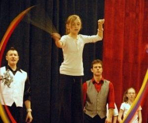 A girl balances on a large green ball while twirling ribbons, with three adults watching nearby.