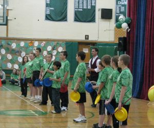 A group of children in green shirts stand in a gym holding colored plates, with an adult and gym decorations in the background.