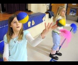 Two girls juggle balls and scarves indoors, one in the foreground concentrating on tossing a yellow and blue ball.