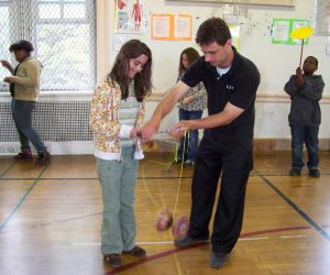 An adult helps a girl use spinning toys in a gym while other children play with different toys in the background.