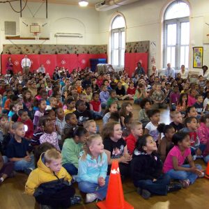 A large group of elementary school children sit on a gym floor, facing forward, with several adults seated in the back.