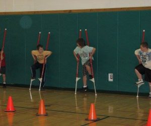 Four children practice walking on stilts in a gymnasium, with safety cones lined up in front of them.
