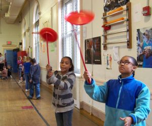 Two children spin red plates on sticks in a school gymnasium, with other kids and posters visible in the background.