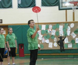 A boy spins a red plate on a stick in a gym as two children watch and art is displayed on the wall behind them.