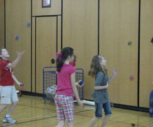 Three children and an adult practice juggling with colorful balls in a gymnasium with wooden walls.