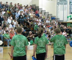 Four students in matching green shirts stand facing a cheering crowd in a school gymnasium decorated with balloons.