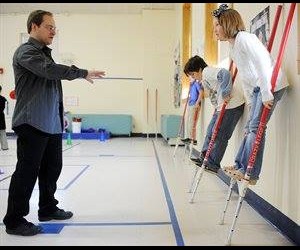 An instructor guides two children balancing on stilts in a gymnasium.