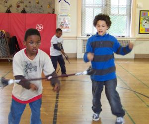 Three children jump rope together in a gymnasium with wooden floors and climbing walls in the background.