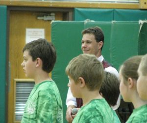 A group of children in green shirts stand in front of a smiling man holding a microphone in a gymnasium.