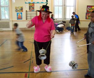 Person in pink shirt, rainbow hat, and pig slippers playing with a diabolo in a gym with children and toys in the background.