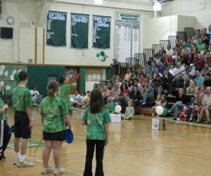 Five children in green shirts stand in a school gymnasium facing an audience seated on bleachers.