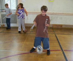 Three children play with spinning tops in a gymnasium with wooden floors and a barred window in the background.