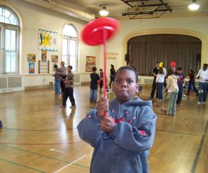 A boy spins a red plate on a stick in a gymnasium with other children participating in similar activities.