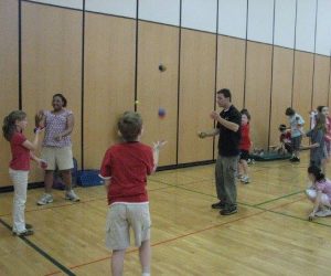 Children and an adult practice juggling and other activities in a gymnasium with wooden floors and paneled walls.