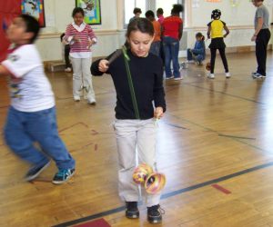 A child plays with a diabolo toy in a gymnasium while other children engage in various activities in the background.