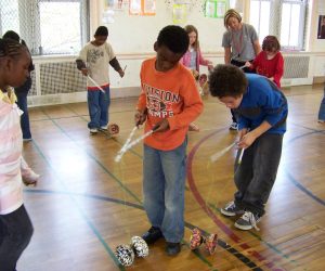 Children practice using Chinese yo-yos in a school gymnasium while an adult supervises in the background.