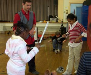 Three children practice spinning diabolos on strings while an adult supervises in a gymnasium.