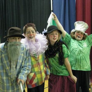 Four children in colorful costumes and makeup pose together indoors, appearing to be in a school gym with curtains behind them.